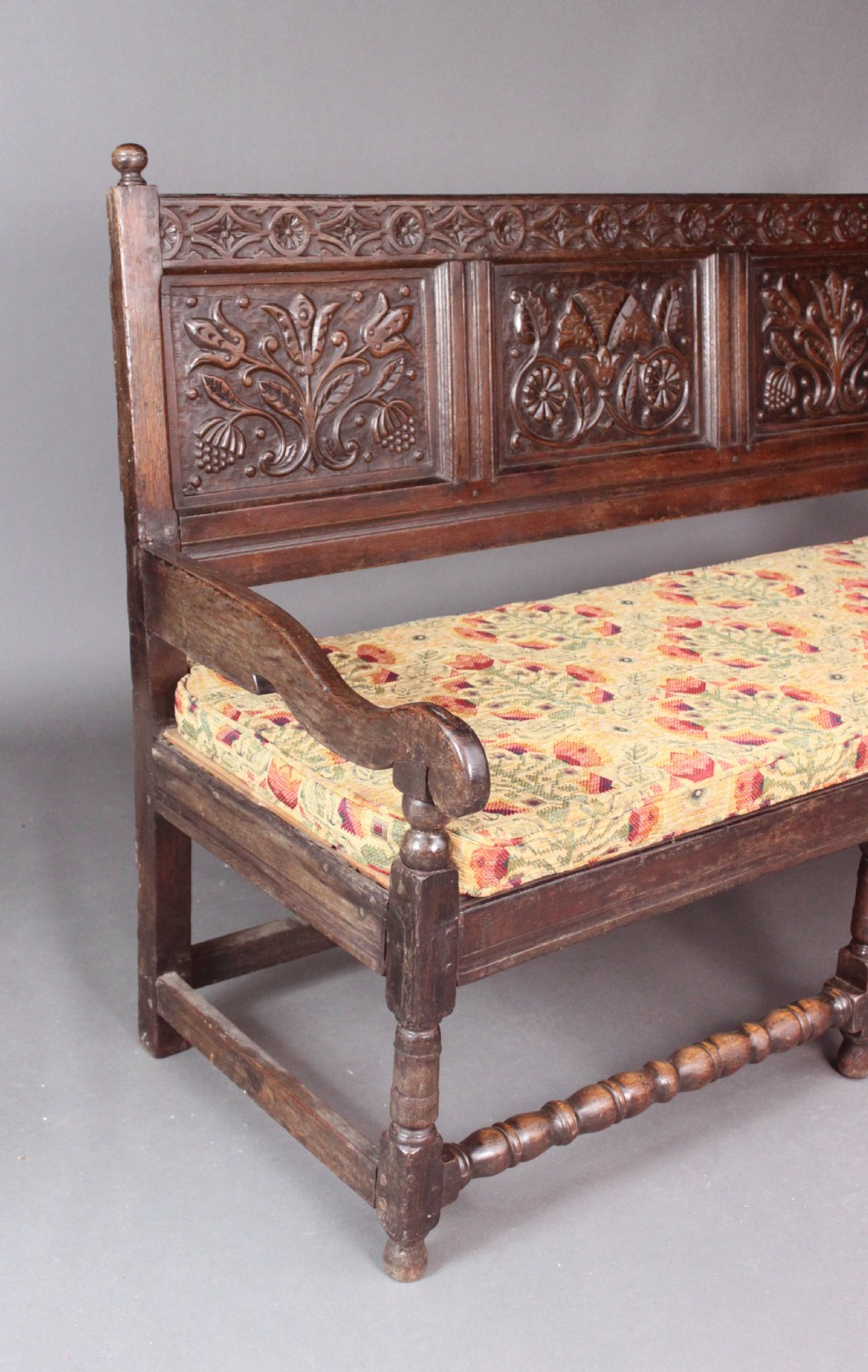A late seventeenth-century dark carved oak settle featuring three carved back panels with floral motifs, turned legs, stretchers, and a floral patterned seat cushion. View III