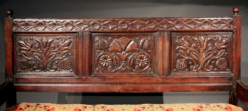 A late seventeenth-century dark carved oak settle featuring three carved back panels with floral motifs, turned legs, stretchers, and a floral patterned seat cushion. View IV