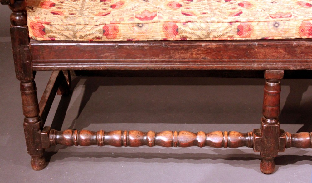 A late seventeenth-century dark carved oak settle featuring three carved back panels with floral motifs, turned legs, stretchers, and a floral patterned seat cushion. View V