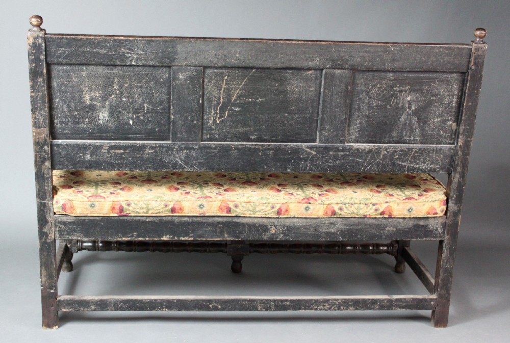 A late seventeenth-century dark carved oak settle featuring three carved back panels with floral motifs, turned legs, stretchers, and a floral patterned seat cushion. View VI
