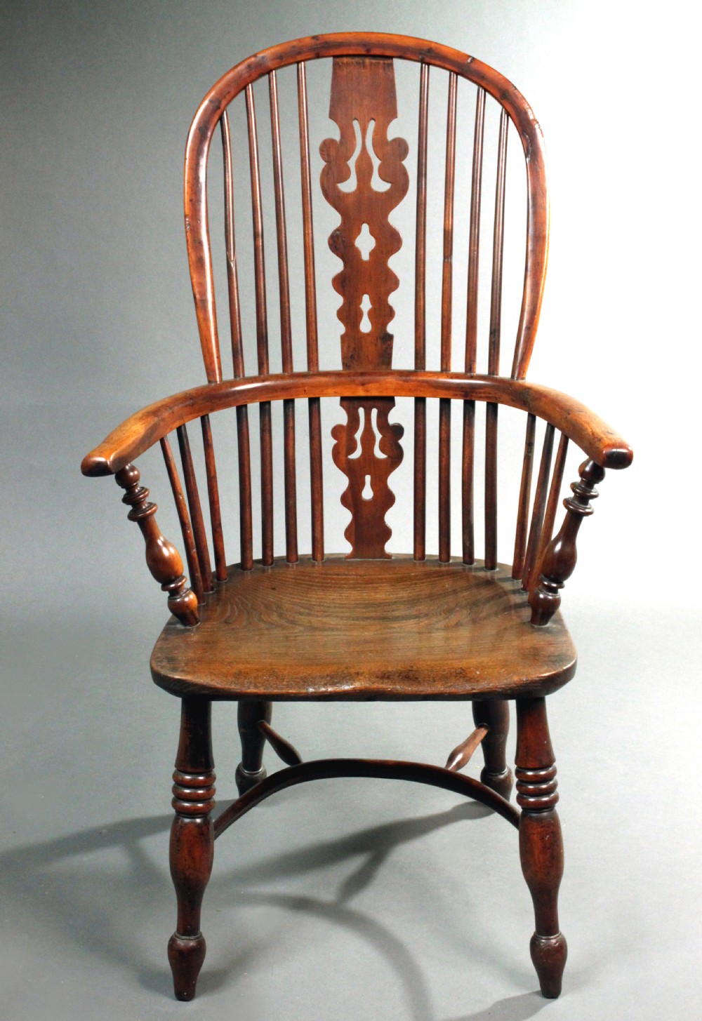 An antique yew wood Windsor armchair featuring a high bow back with vertical spindles, a pierced central splat, turned arm supports, and a shaped seat.