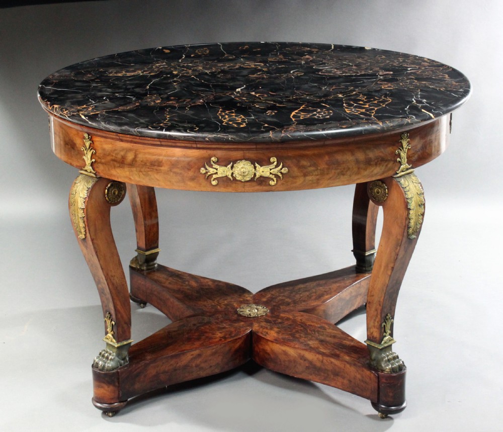 A circular French Empire centre table featuring a black veined marble top, mahogany apron with gilt bronze mounts, and curved legs on a cruciform base.