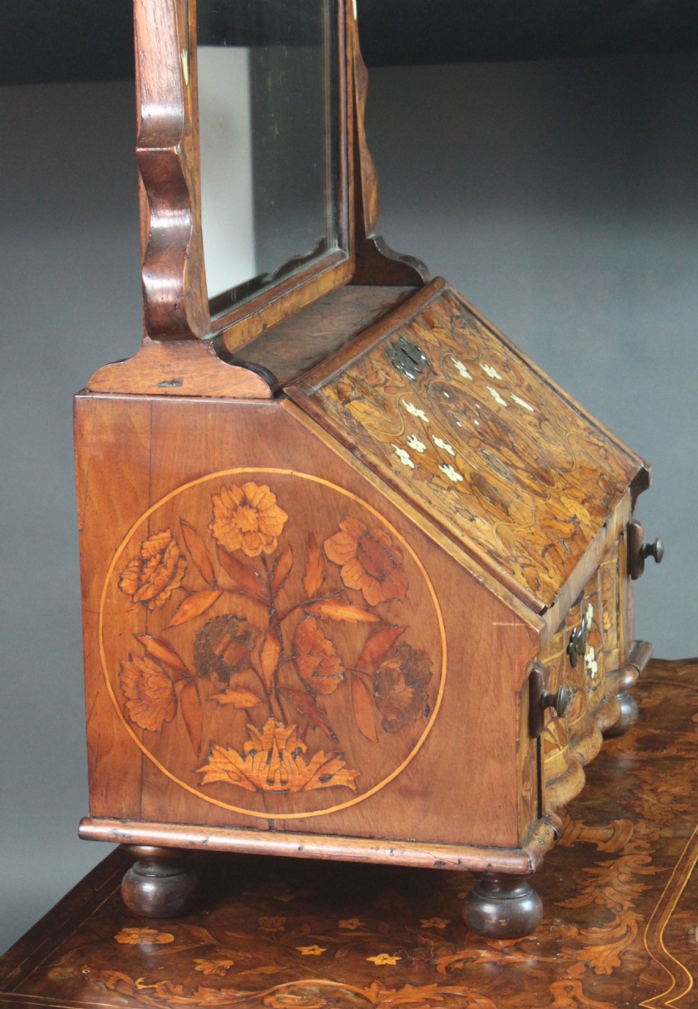 An antique Dutch walnut toilet mirror featuring detailed floral marquetry inlay, an arched frame, and a slant-front drawer base supported by bun feet. View VII