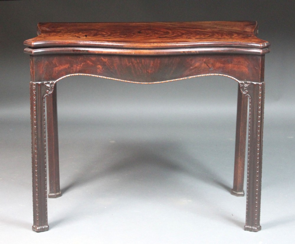An 18th-century Chippendale style mahogany card table featuring a serpentine fold-over top, carved apron edge, and square moulded legs.