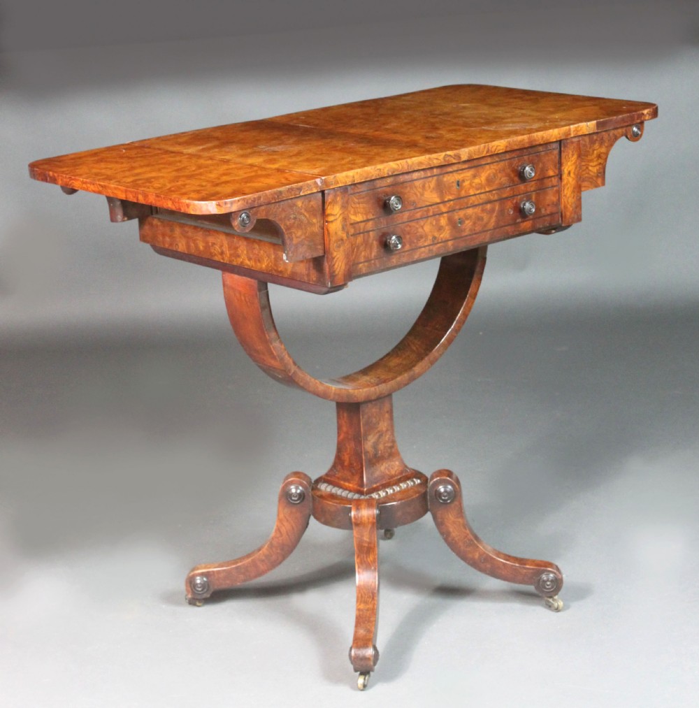 A Regency period burr ash work table featuring drop leaves, two small drawers with round knobs, a U-shaped pedestal support, and four splayed legs with brass castors.