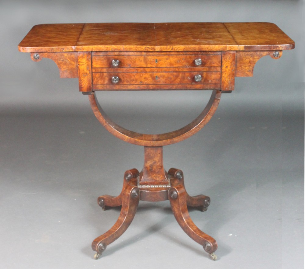 A Regency period burr ash work table featuring drop leaves, two small drawers with round knobs, a U-shaped pedestal support, and four splayed legs with brass castors. View II