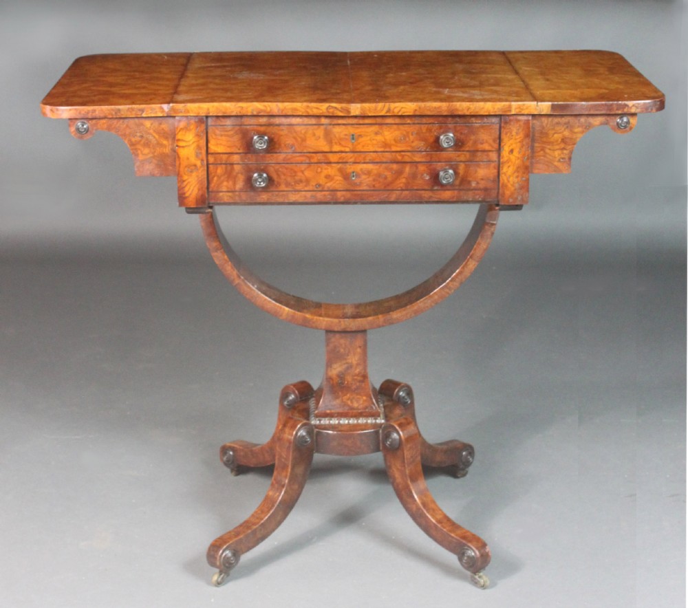 A Regency period burr ash work table featuring drop leaves, two small drawers with round knobs, a U-shaped pedestal support, and four splayed legs with brass castors. View III
