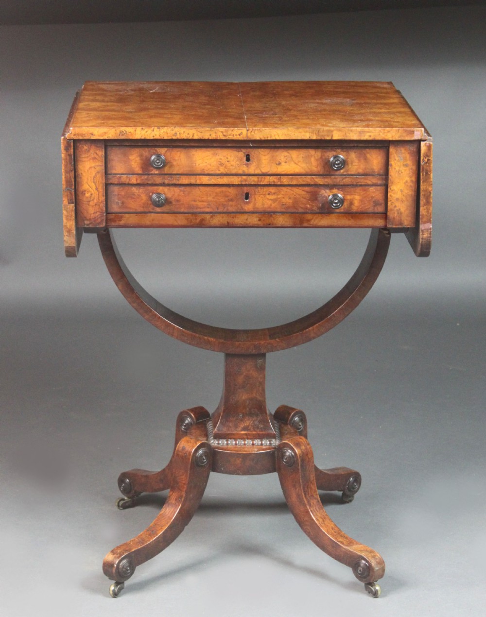 A Regency period burr ash work table featuring drop leaves, two small drawers with round knobs, a U-shaped pedestal support, and four splayed legs with brass castors. View V