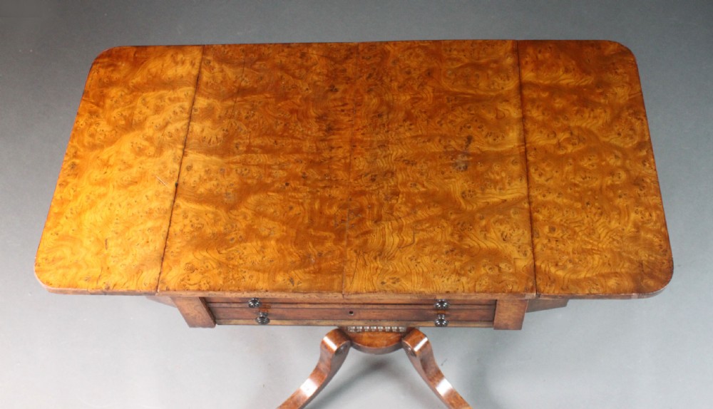 A Regency period burr ash work table featuring drop leaves, two small drawers with round knobs, a U-shaped pedestal support, and four splayed legs with brass castors. View VI