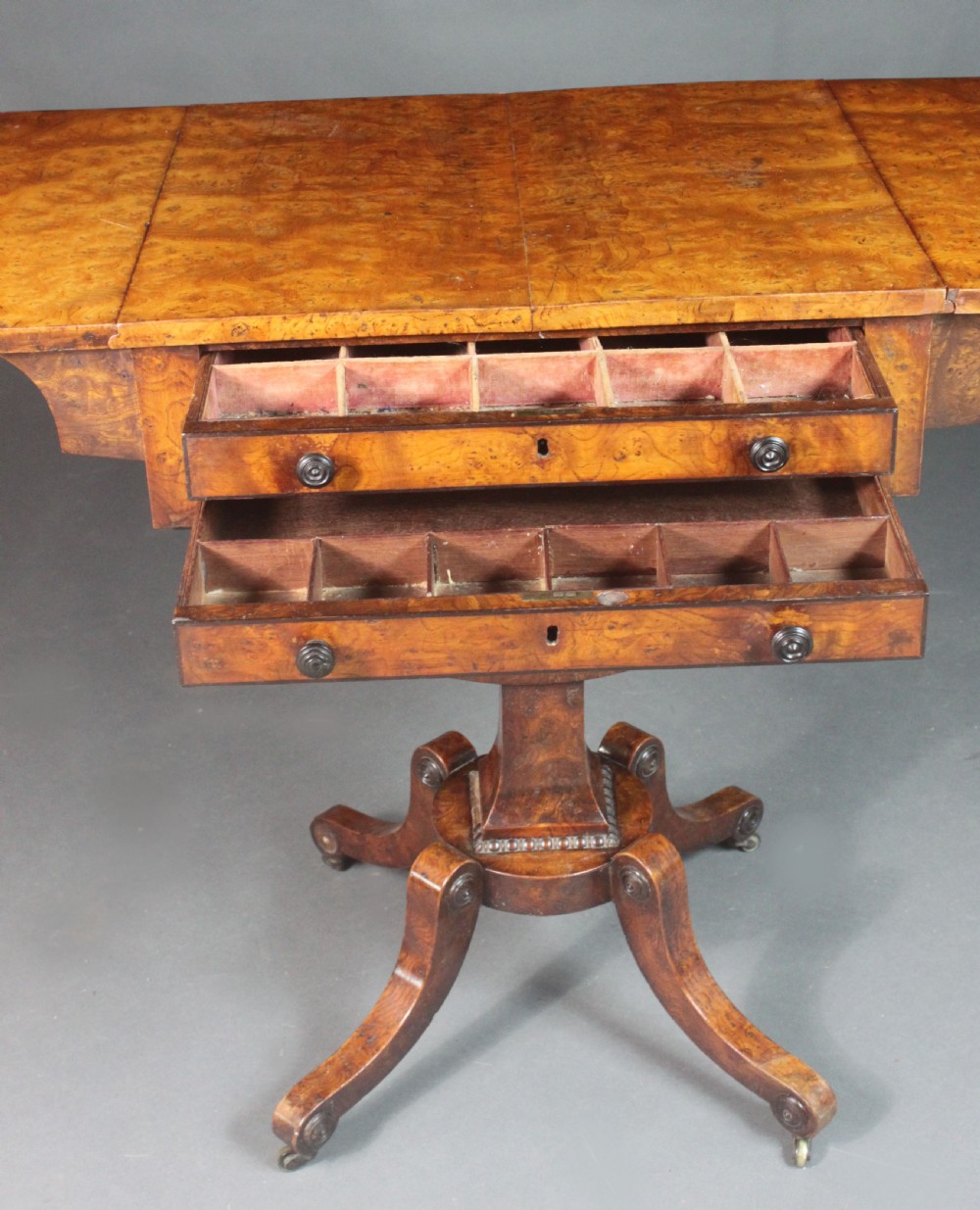 A Regency period burr ash work table featuring drop leaves, two small drawers with round knobs, a U-shaped pedestal support, and four splayed legs with brass castors. View VII