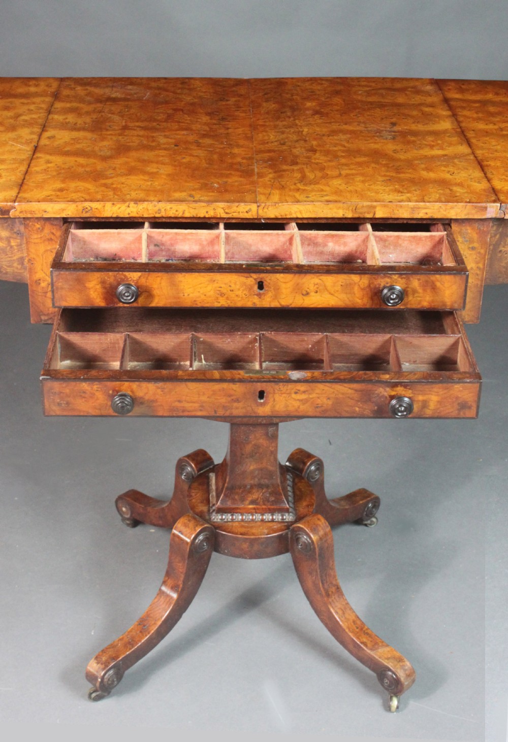 A Regency period burr ash work table featuring drop leaves, two small drawers with round knobs, a U-shaped pedestal support, and four splayed legs with brass castors. View VIII