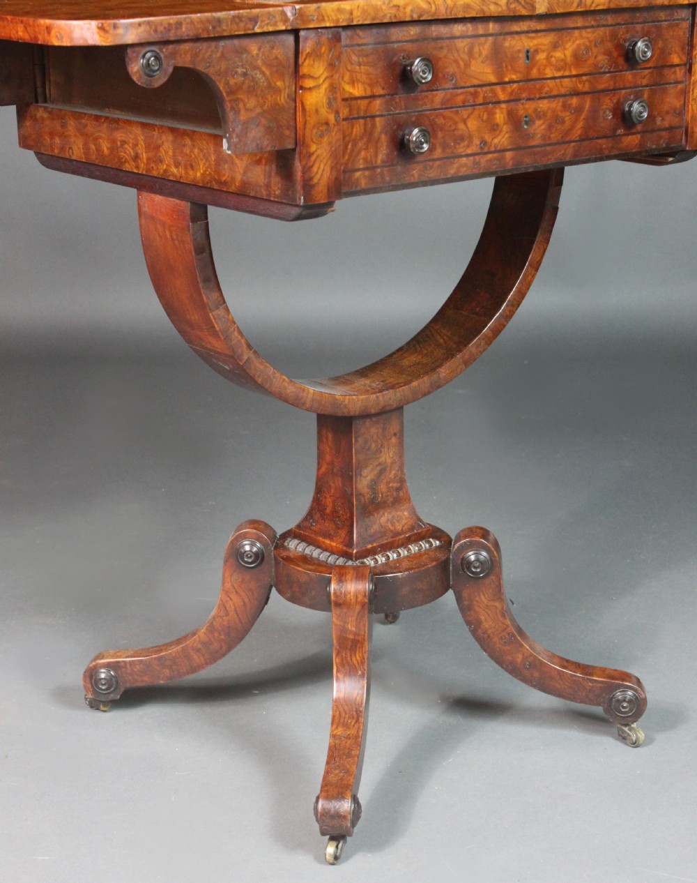 A Regency period burr ash work table featuring drop leaves, two small drawers with round knobs, a U-shaped pedestal support, and four splayed legs with brass castors. View IX