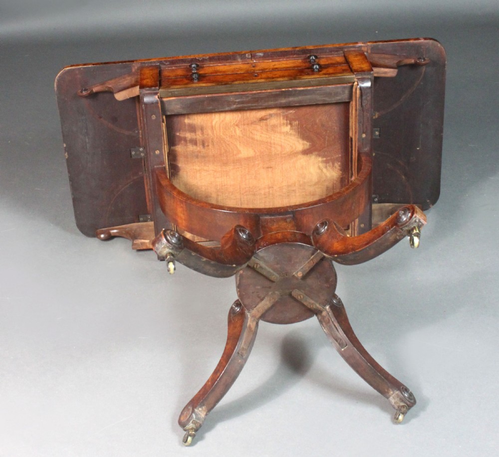 A Regency period burr ash work table featuring drop leaves, two small drawers with round knobs, a U-shaped pedestal support, and four splayed legs with brass castors. View X