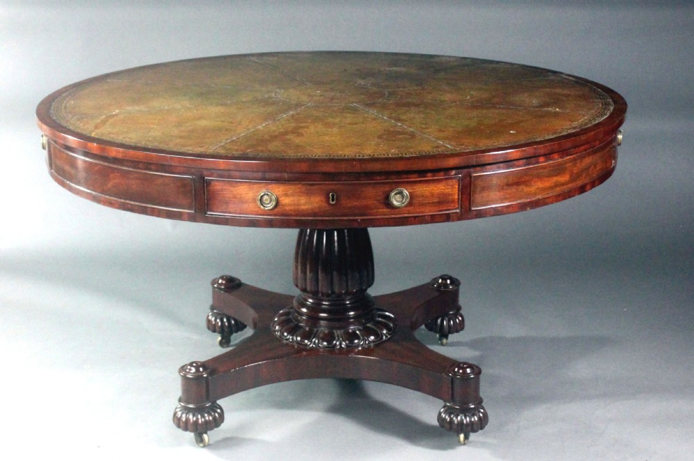 A circular Georgian mahogany drum table featuring a green leather inset top with gilt tooling, frieze drawers with brass handles, and a carved pedestal base resting on four splayed legs with castors.