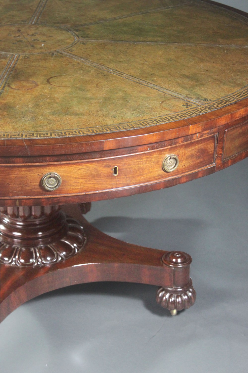 A circular Georgian mahogany drum table featuring a green leather inset top with gilt tooling, frieze drawers with brass handles, and a carved pedestal base resting on four splayed legs with castors. View VI