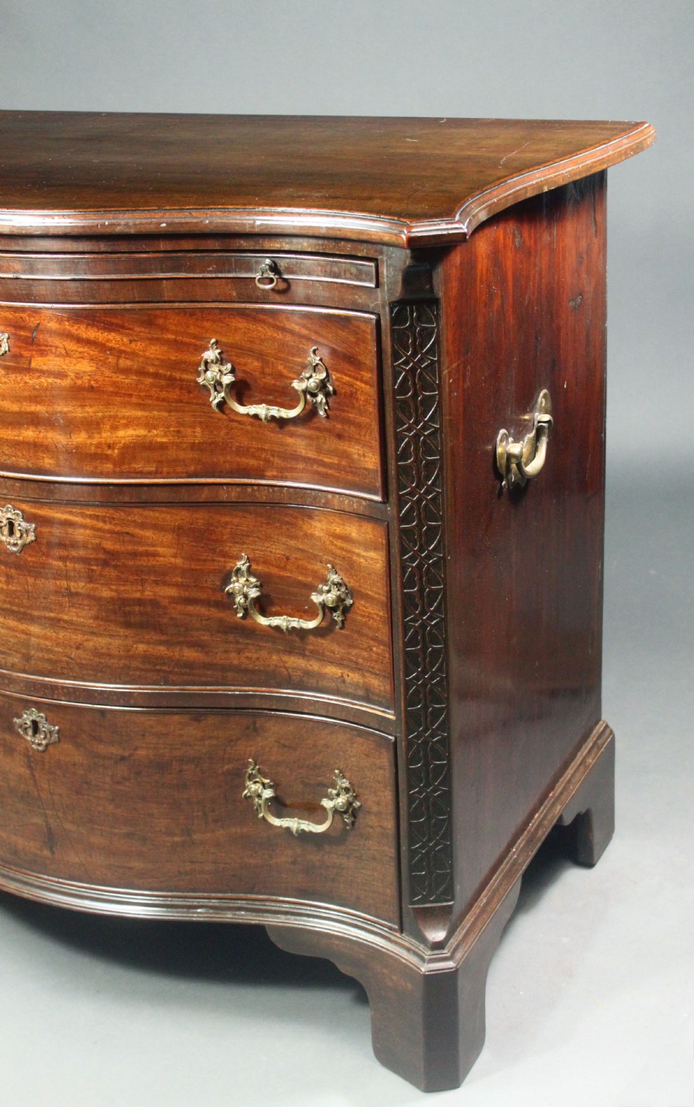 An eighteenth-century George III mahogany chest of drawers featuring a serpentine front, carved blind fretwork, brass handles, and bracket feet. View V