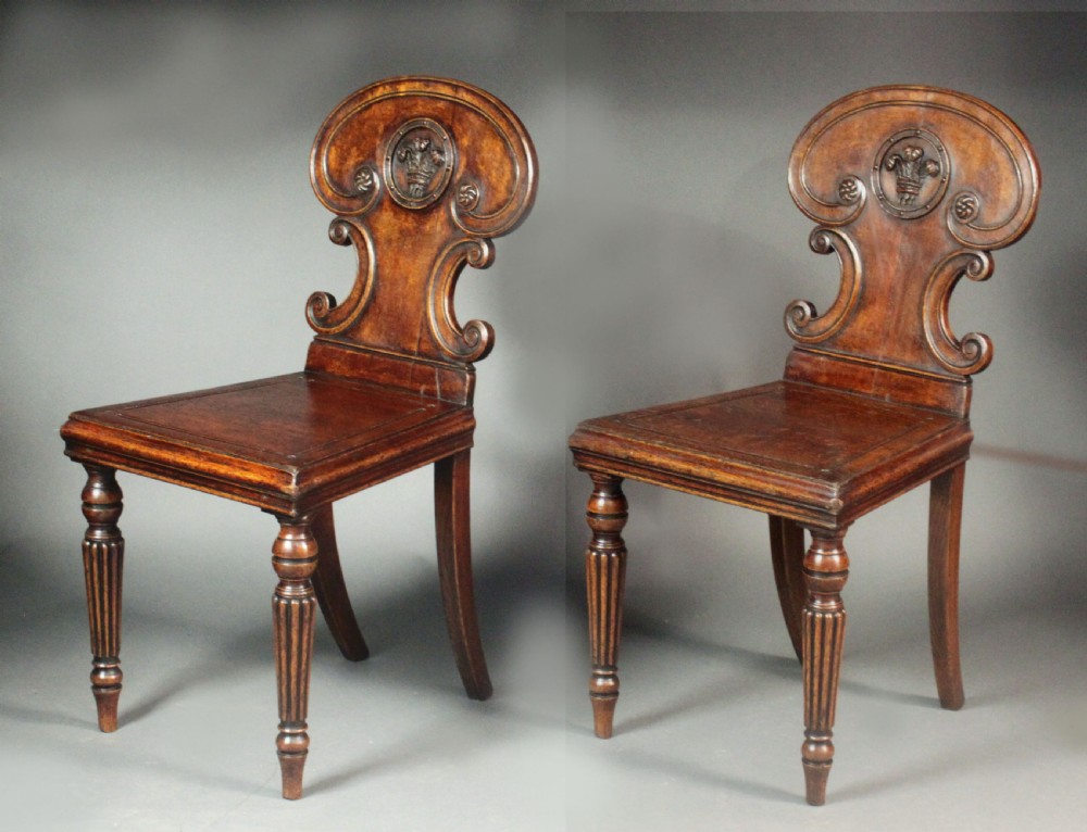 A pair of Regency oak hall chairs featuring shaped scroll backrests with central carved medallions, solid seats, and turned fluted front legs.