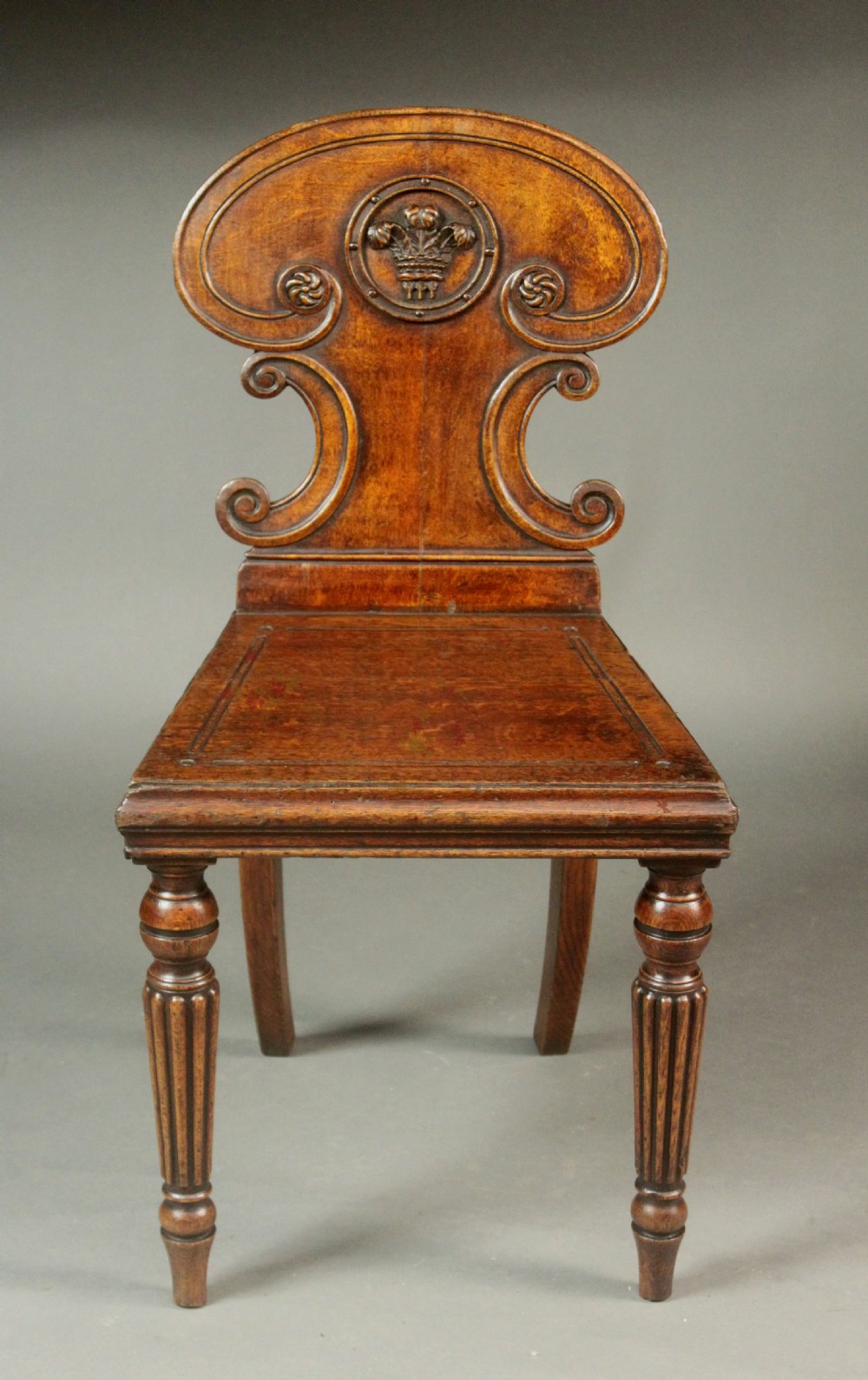 A pair of Regency oak hall chairs featuring shaped scroll backrests with central carved medallions, solid seats, and turned fluted front legs. View V