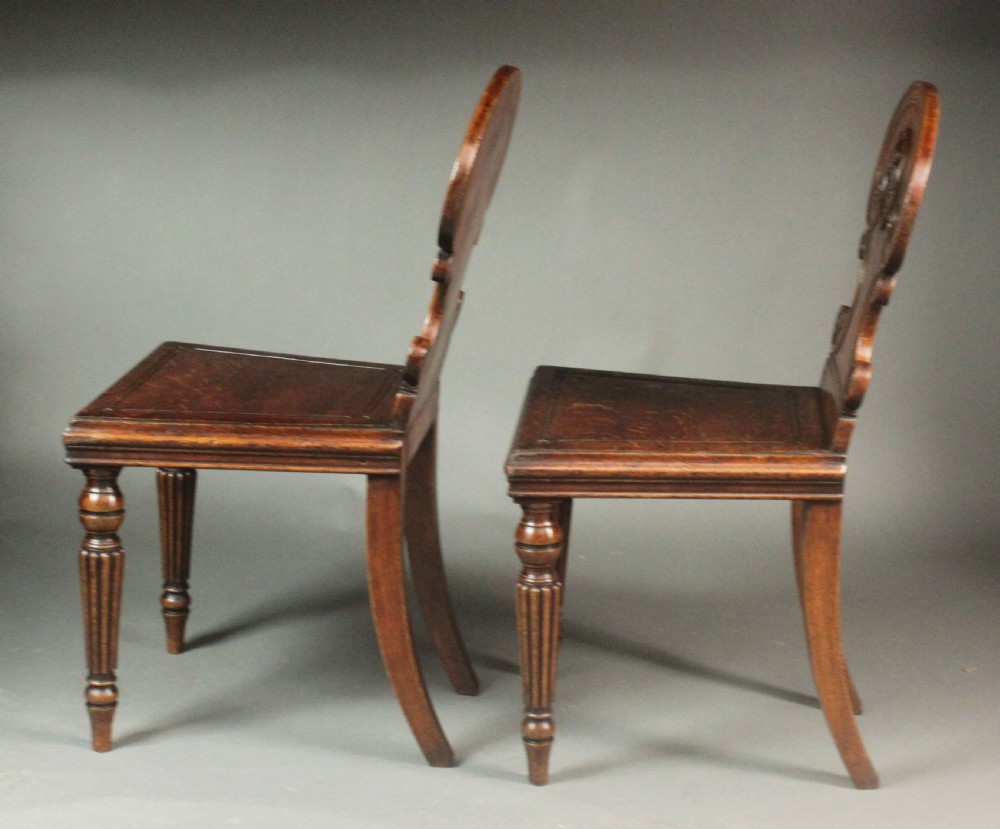 A pair of Regency oak hall chairs featuring shaped scroll backrests with central carved medallions, solid seats, and turned fluted front legs. View VI