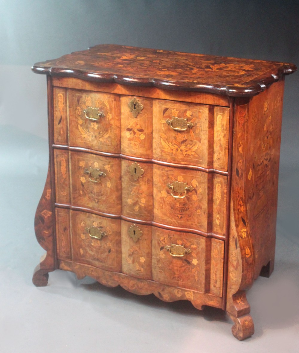 An antique Dutch marquetry chest of drawers featuring three drawers, floral wood inlay, shaped sides, and brass drop handles.