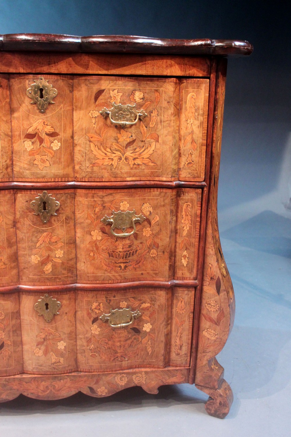 An antique Dutch marquetry chest of drawers featuring three drawers, floral wood inlay, shaped sides, and brass drop handles. View VI