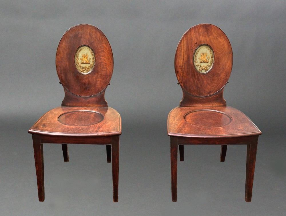 A pair of George III mahogany hall chairs featuring oval backrests with central painted armorial crests, solid timber seats, and square tapered legs.