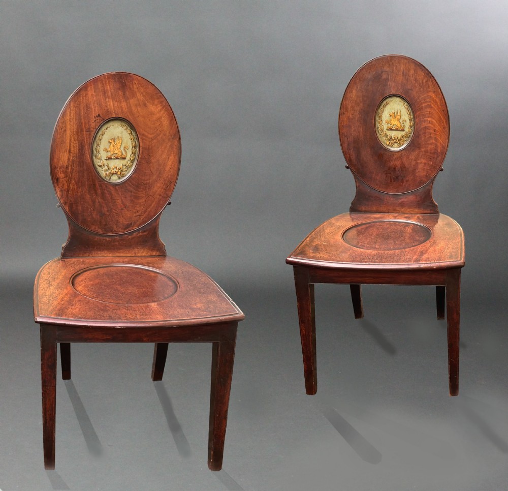 A pair of George III mahogany hall chairs featuring oval backrests with central painted armorial crests, solid timber seats, and square tapered legs. View II