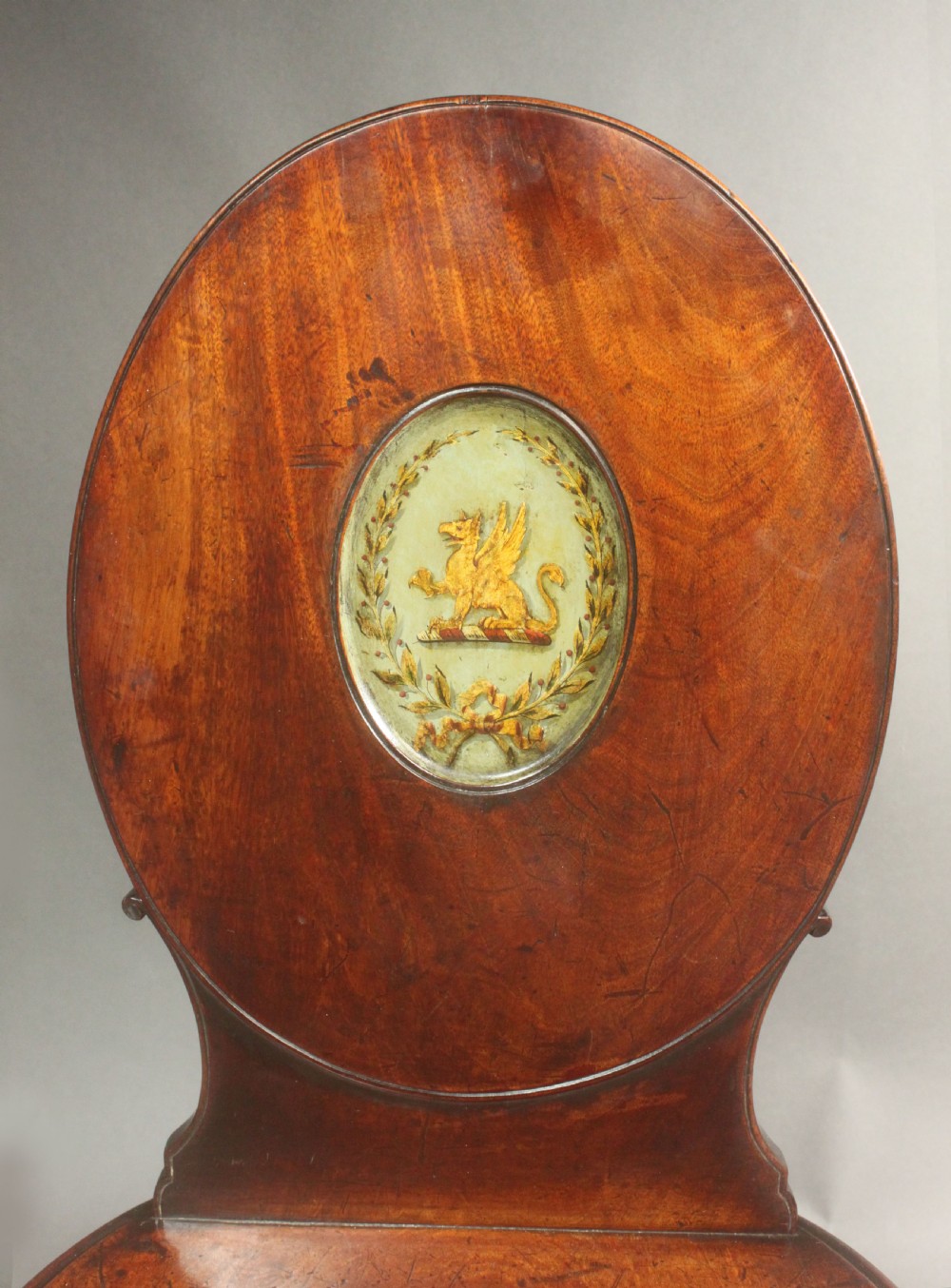 A pair of George III mahogany hall chairs featuring oval backrests with central painted armorial crests, solid timber seats, and square tapered legs. View IV