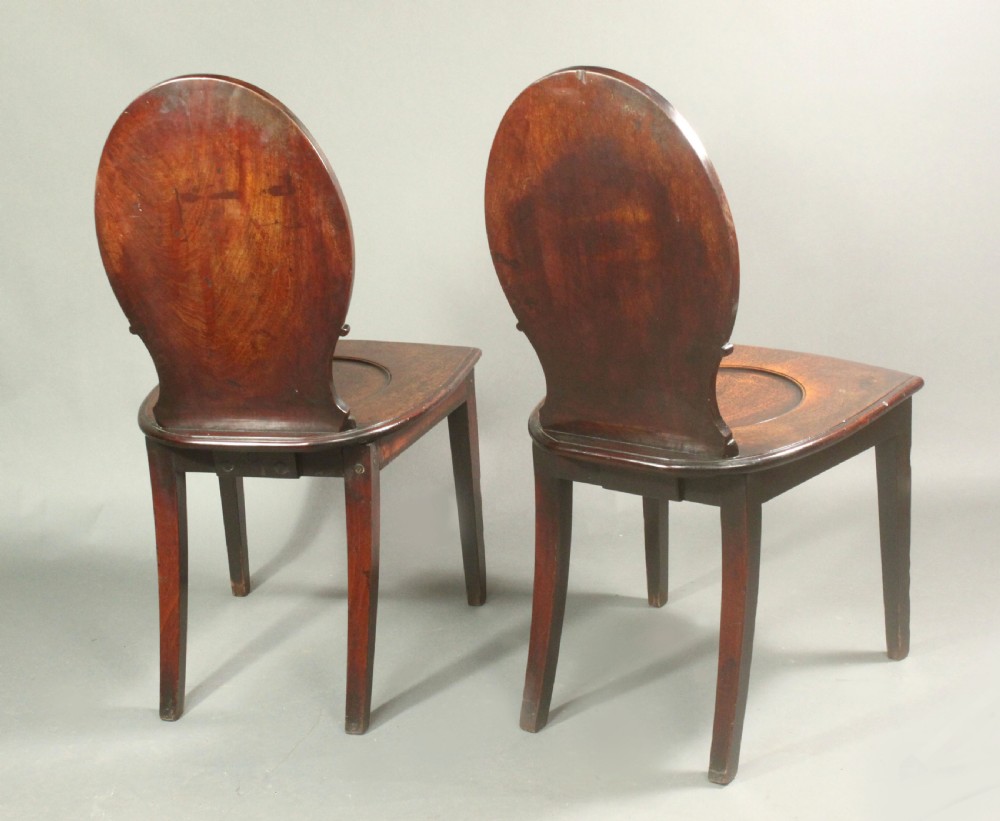 A pair of George III mahogany hall chairs featuring oval backrests with central painted armorial crests, solid timber seats, and square tapered legs. View VI