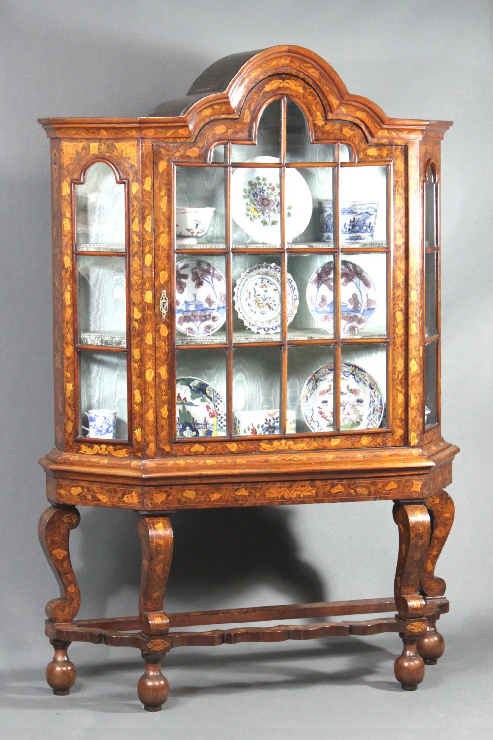 An antique Dutch marquetry glazed cabinet featuring intricate floral wood inlays, glazed doors, an arched pediment, and shaped legs with a stretcher base.