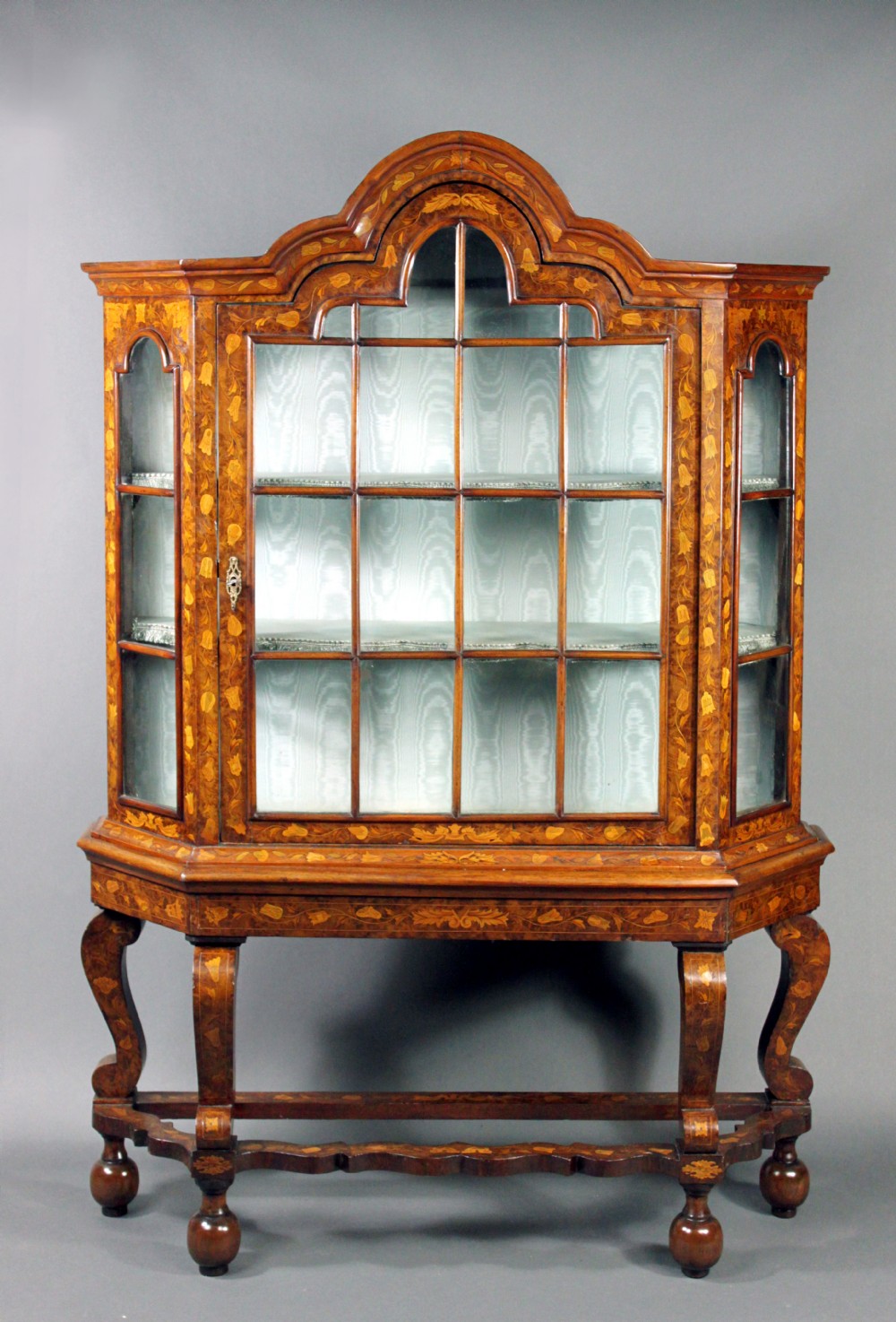 An antique Dutch marquetry glazed cabinet featuring intricate floral wood inlays, glazed doors, an arched pediment, and shaped legs with a stretcher base. View II