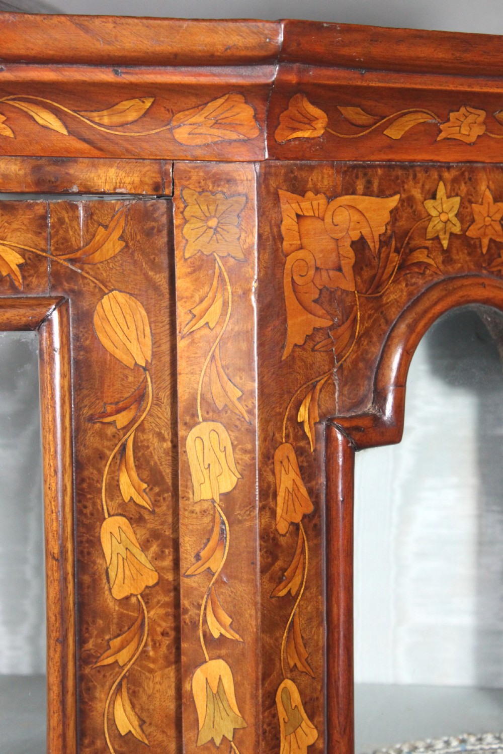 An antique Dutch marquetry glazed cabinet featuring intricate floral wood inlays, glazed doors, an arched pediment, and shaped legs with a stretcher base. View VI