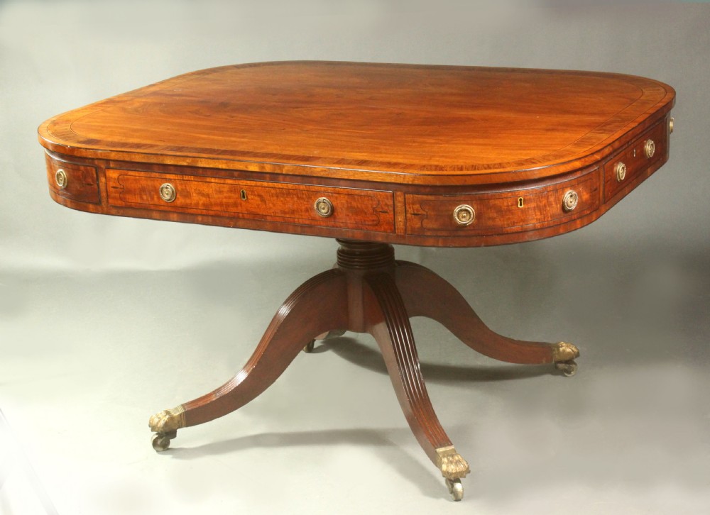 A Regency mahogany centre table featuring a rounded rectangular top, crossbanded border, drawer frieze with brass ring handles, and a reeded quadruped pedestal base with brass lion paw feet on castors.