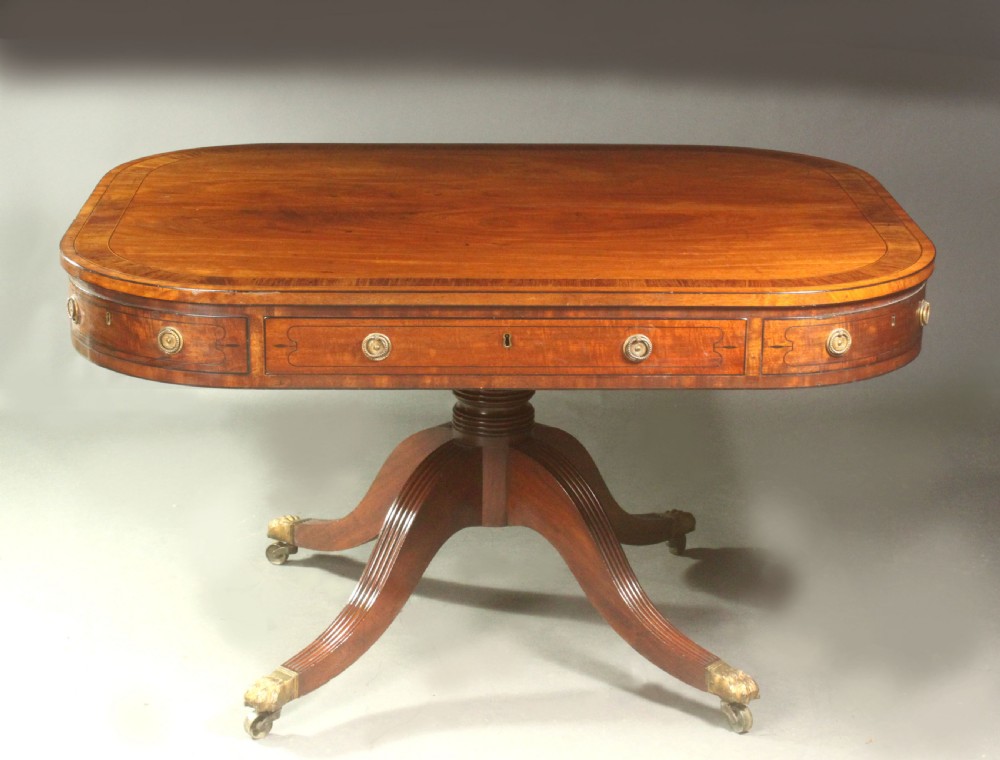 A Regency mahogany centre table featuring a rounded rectangular top, crossbanded border, drawer frieze with brass ring handles, and a reeded quadruped pedestal base with brass lion paw feet on castors. View II