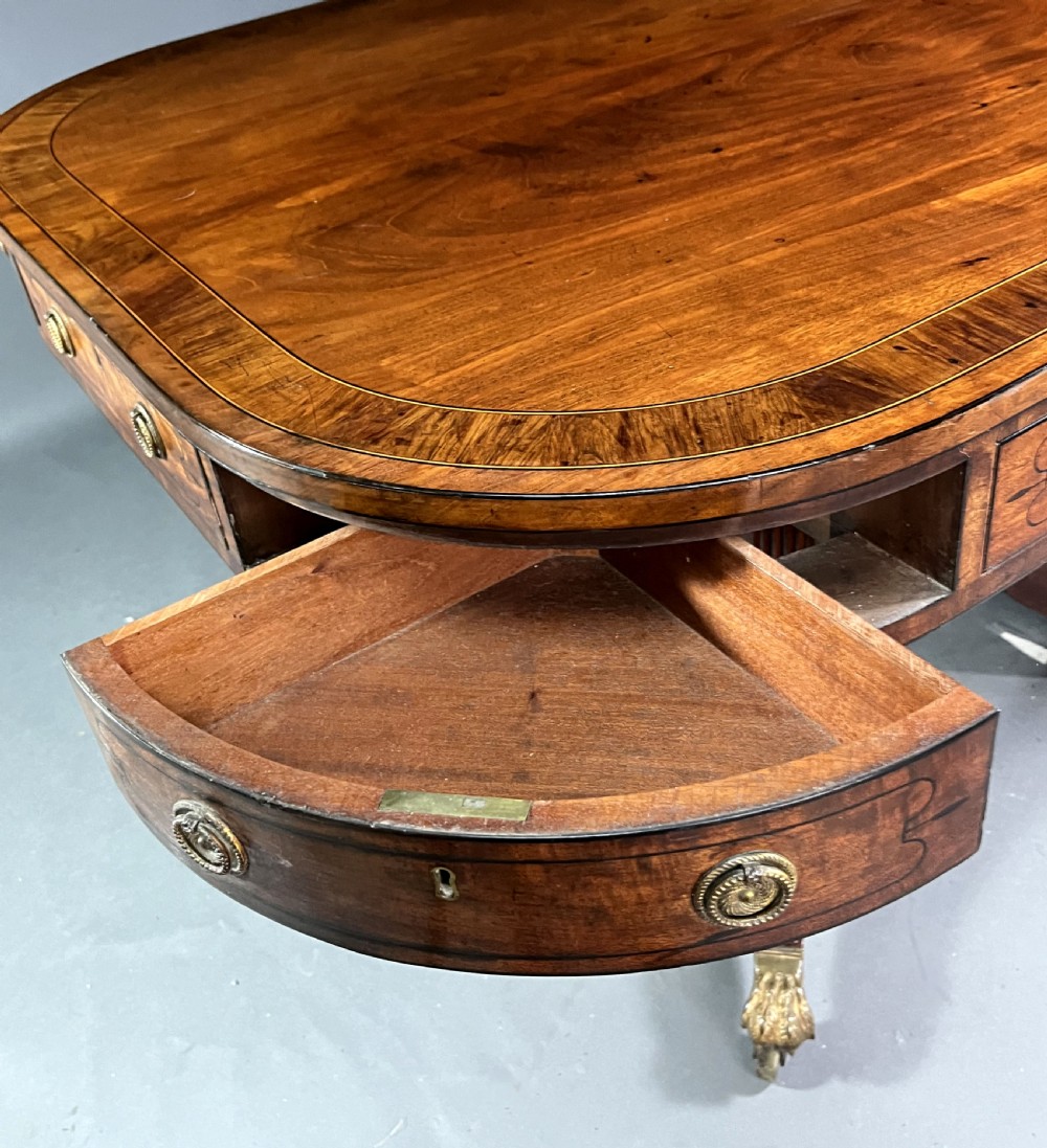 A Regency mahogany centre table featuring a rounded rectangular top, crossbanded border, drawer frieze with brass ring handles, and a reeded quadruped pedestal base with brass lion paw feet on castors. View III
