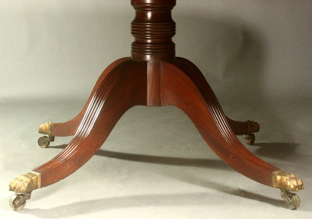 A Regency mahogany centre table featuring a rounded rectangular top, crossbanded border, drawer frieze with brass ring handles, and a reeded quadruped pedestal base with brass lion paw feet on castors. View IV