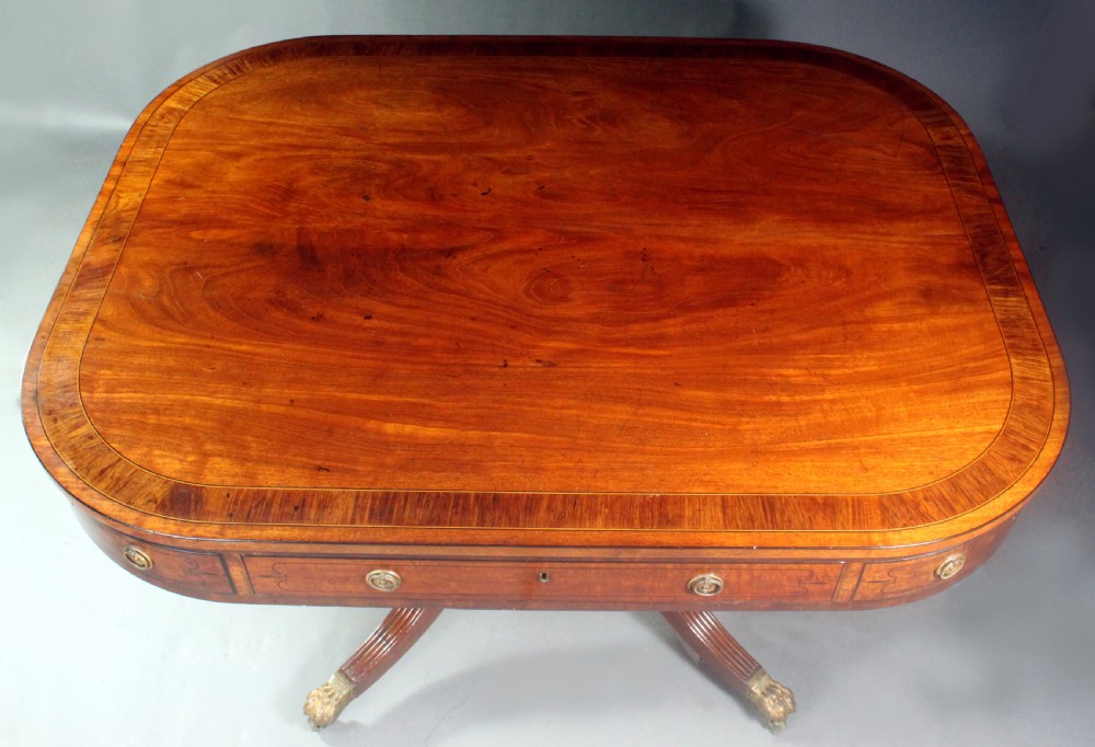 A Regency mahogany centre table featuring a rounded rectangular top, crossbanded border, drawer frieze with brass ring handles, and a reeded quadruped pedestal base with brass lion paw feet on castors. View VI