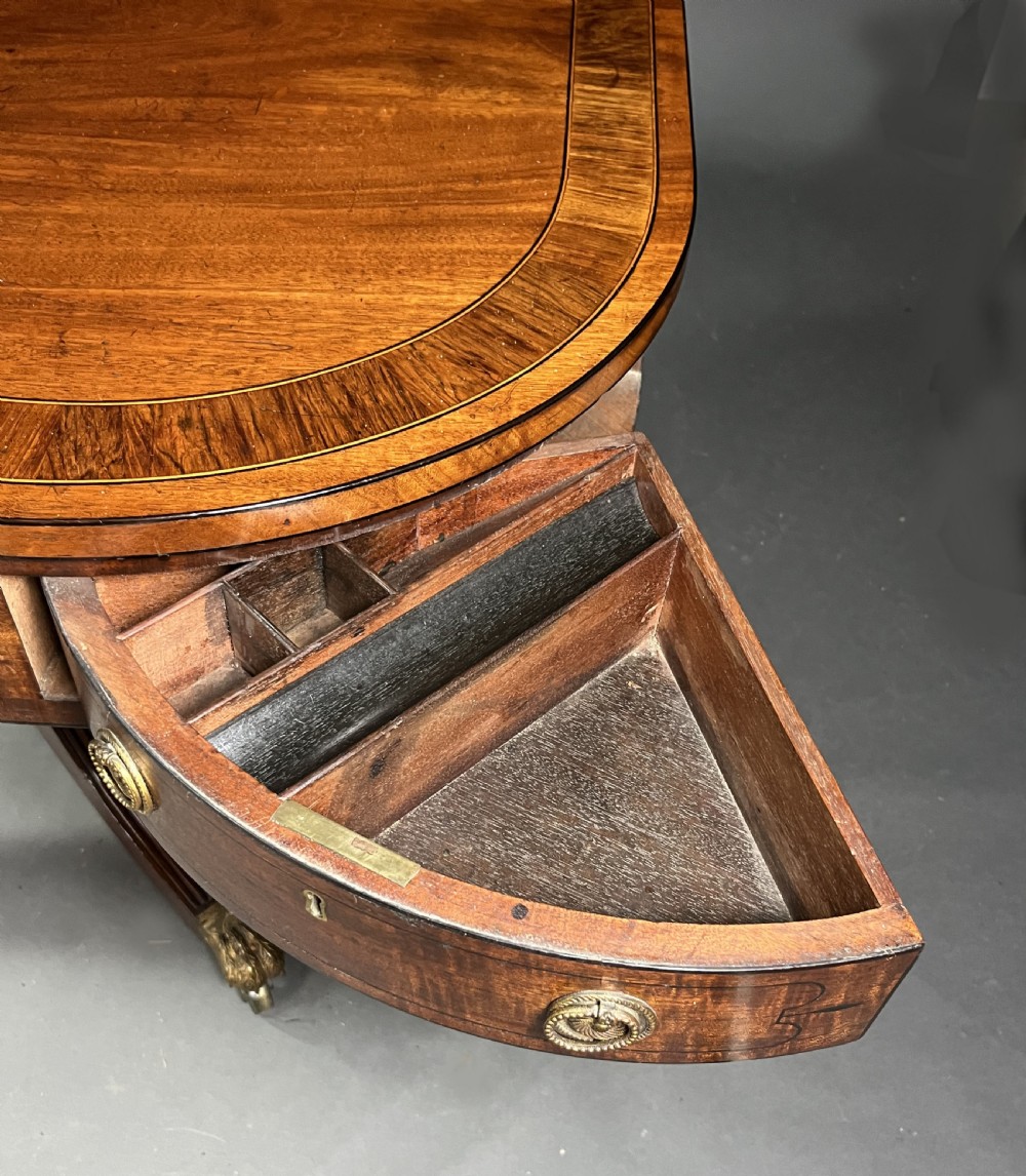 A Regency mahogany centre table featuring a rounded rectangular top, crossbanded border, drawer frieze with brass ring handles, and a reeded quadruped pedestal base with brass lion paw feet on castors. View VIII