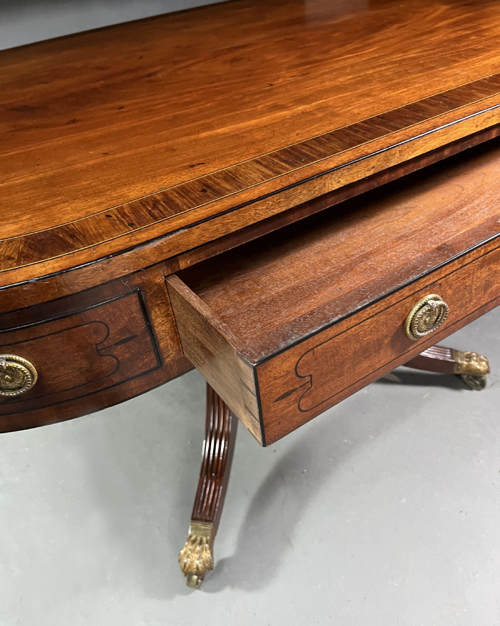A Regency mahogany centre table featuring a rounded rectangular top, crossbanded border, drawer frieze with brass ring handles, and a reeded quadruped pedestal base with brass lion paw feet on castors. View X