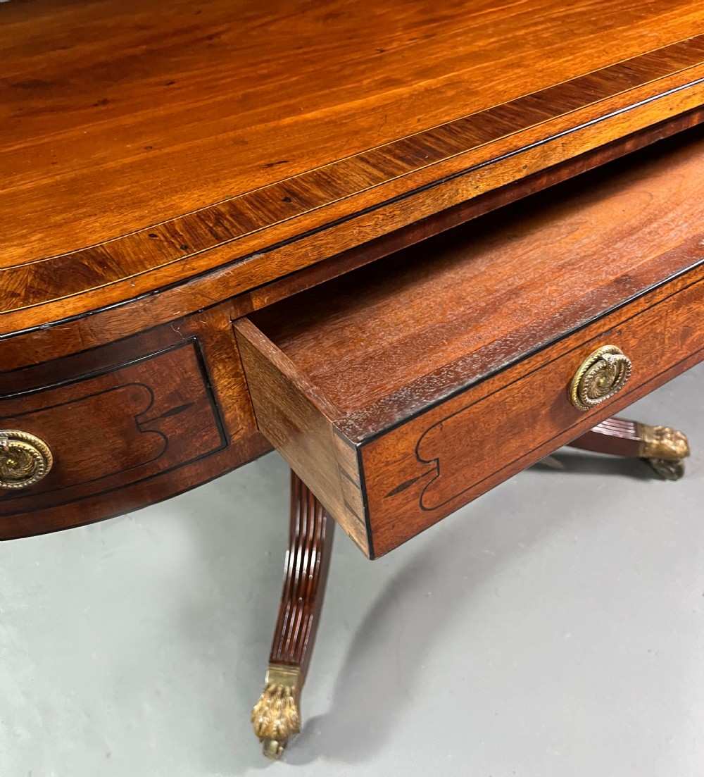 A Regency mahogany centre table featuring a rounded rectangular top, crossbanded border, drawer frieze with brass ring handles, and a reeded quadruped pedestal base with brass lion paw feet on castors. View XII