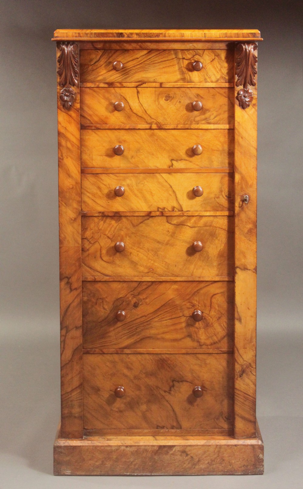 A tall Victorian walnut Wellington chest featuring seven graduated drawers with turned wooden knobs, carved upper corbels, and a right-hand hinged locking flap.