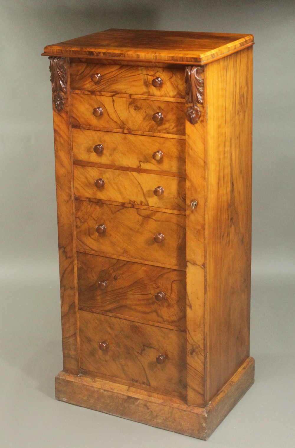A tall Victorian walnut Wellington chest featuring seven graduated drawers with turned wooden knobs, carved upper corbels, and a right-hand hinged locking flap. View II