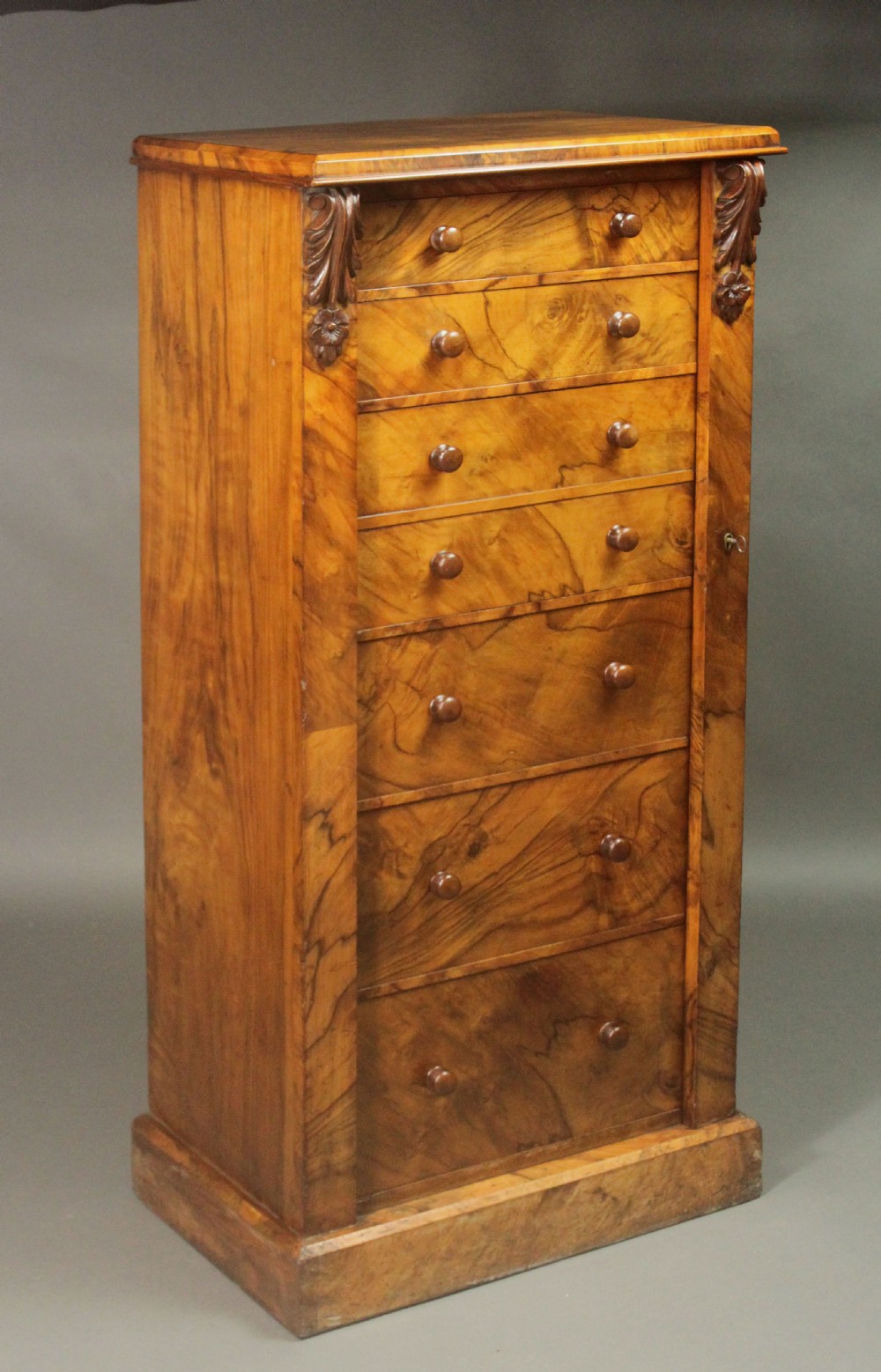 A tall Victorian walnut Wellington chest featuring seven graduated drawers with turned wooden knobs, carved upper corbels, and a right-hand hinged locking flap. View III