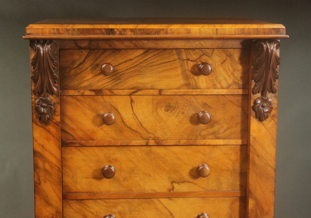 A tall Victorian walnut Wellington chest featuring seven graduated drawers with turned wooden knobs, carved upper corbels, and a right-hand hinged locking flap. View V