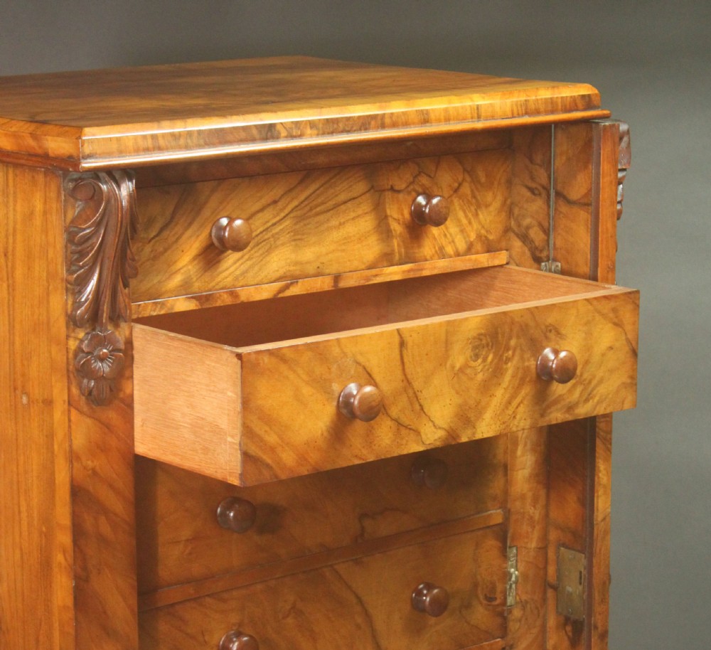 A tall Victorian walnut Wellington chest featuring seven graduated drawers with turned wooden knobs, carved upper corbels, and a right-hand hinged locking flap. View VII