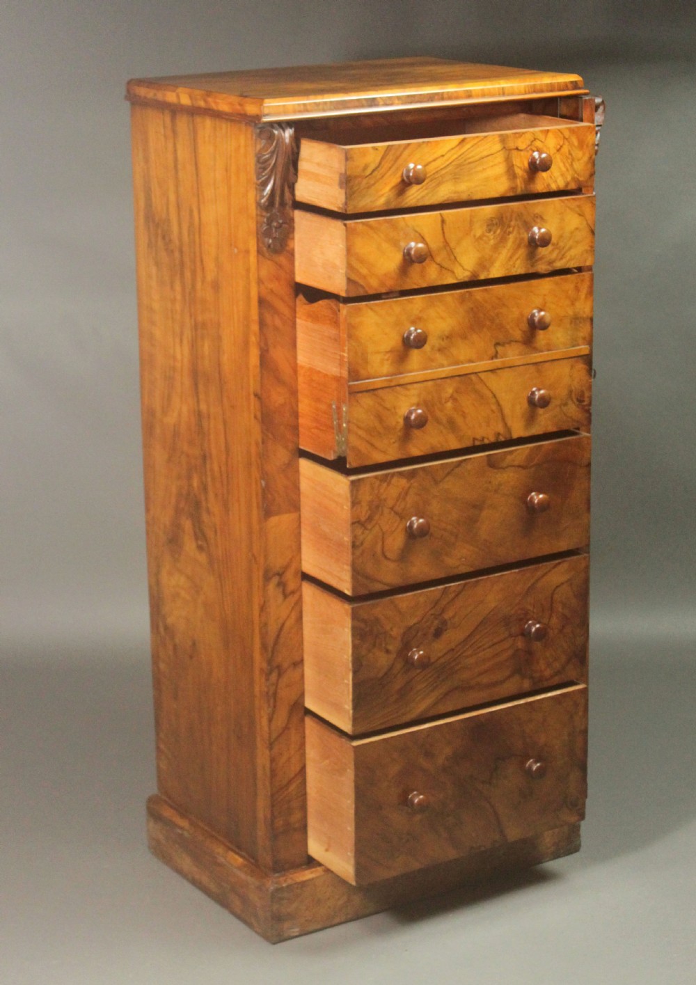 A tall Victorian walnut Wellington chest featuring seven graduated drawers with turned wooden knobs, carved upper corbels, and a right-hand hinged locking flap. View X