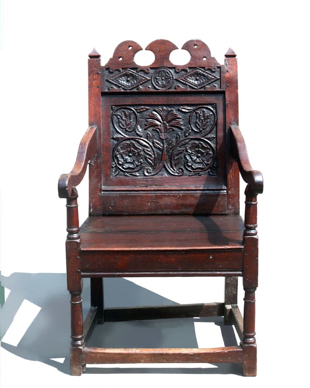 A dark oak wainscot armchair featuring a carved botanical back panel, turned front legs, flat wooden seat, and a crest rail with carved geometric details.