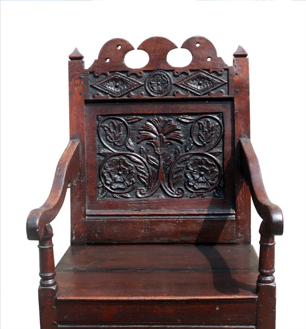 A dark oak wainscot armchair featuring a carved botanical back panel, turned front legs, flat wooden seat, and a crest rail with carved geometric details. View III