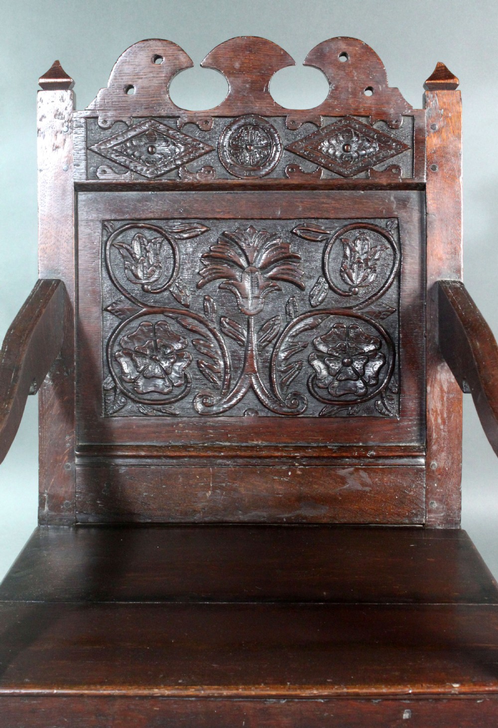A dark oak wainscot armchair featuring a carved botanical back panel, turned front legs, flat wooden seat, and a crest rail with carved geometric details. View IV