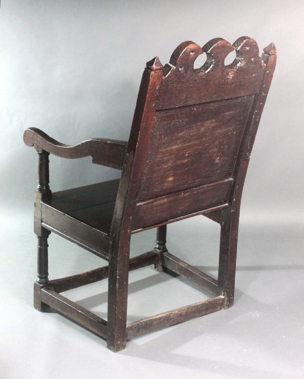 A dark oak wainscot armchair featuring a carved botanical back panel, turned front legs, flat wooden seat, and a crest rail with carved geometric details. View V